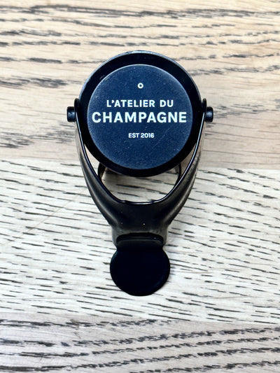 Black champagne stopper with "L'Atelier du Champagne" and "Est 2016" text on a wooden surface.